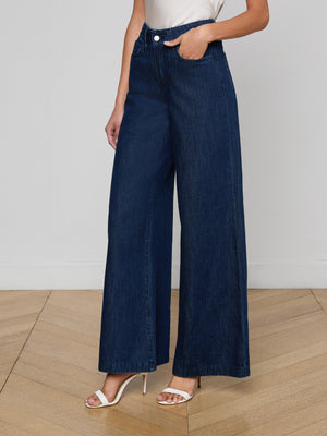 A person wears the Pacey Wide-Leg Jean in a dark wash, paired with a white sleeveless top and white heeled sandals. The photo shows only the lower half of their body on a wooden floor against a plain white wall.