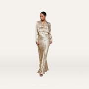 A woman stands indoors on a wooden floor, exuding minimalist sophistication in the cream Ryker Silk Maxi Skirt and a matching patterned blouse, her hair pulled back as she gazes to the side with one hand in her pocket.