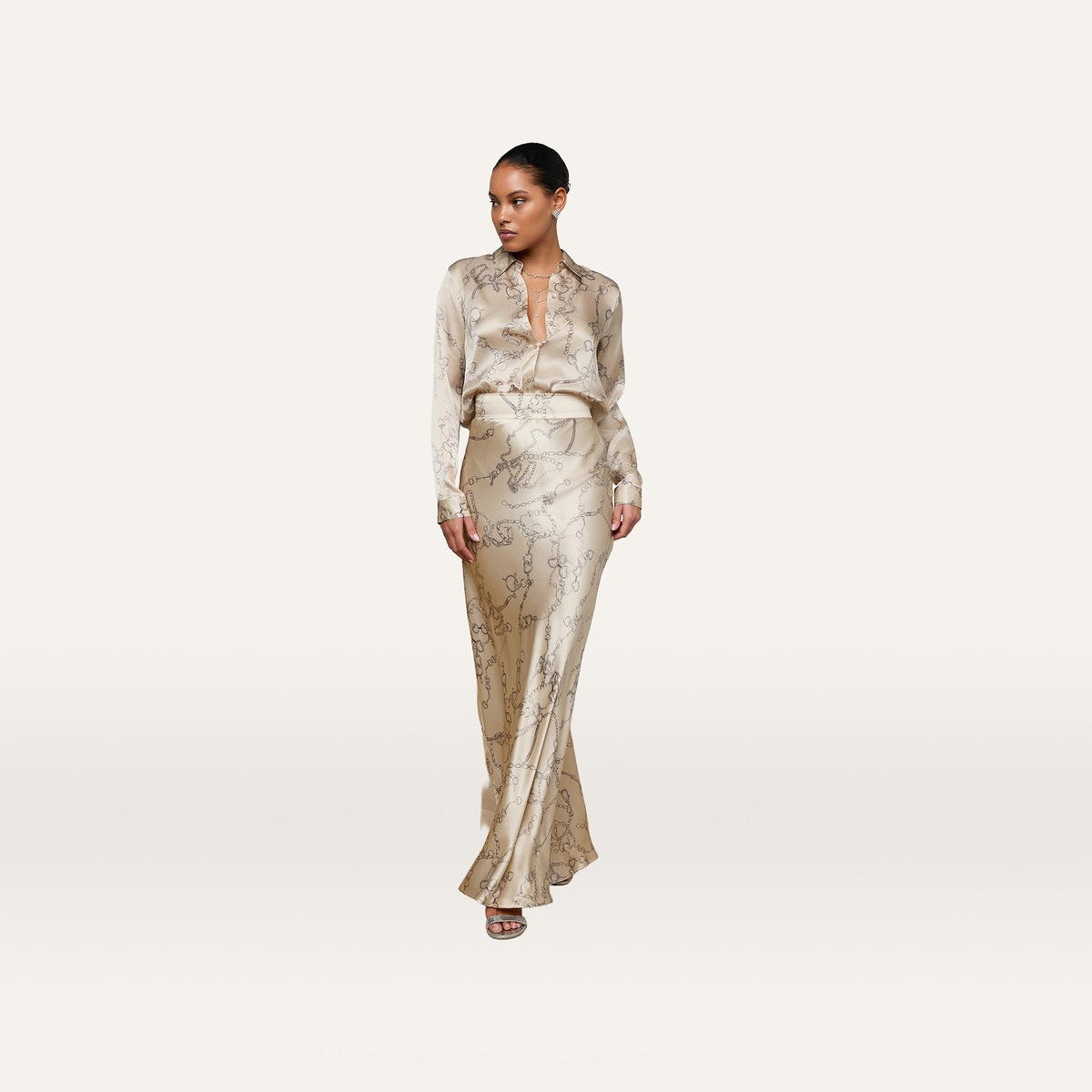 A woman stands indoors on a wooden floor, exuding minimalist sophistication in the cream Ryker Silk Maxi Skirt and a matching patterned blouse, her hair pulled back as she gazes to the side with one hand in her pocket.
