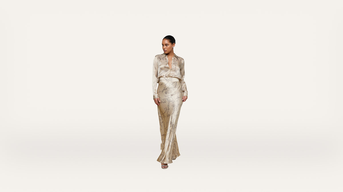 A woman stands confidently wearing a silky, light-colored, long-sleeve blouse and matching flowing skirt with a subtle pattern, against a plain, light background.