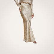 A person wears the Ryker Silk Maxi Skirt in shiny beige with a subtle horsebit chain print, paired with a matching long-sleeve top and open-toe sandals, creating a look of minimalist sophistication on a light wood floor.