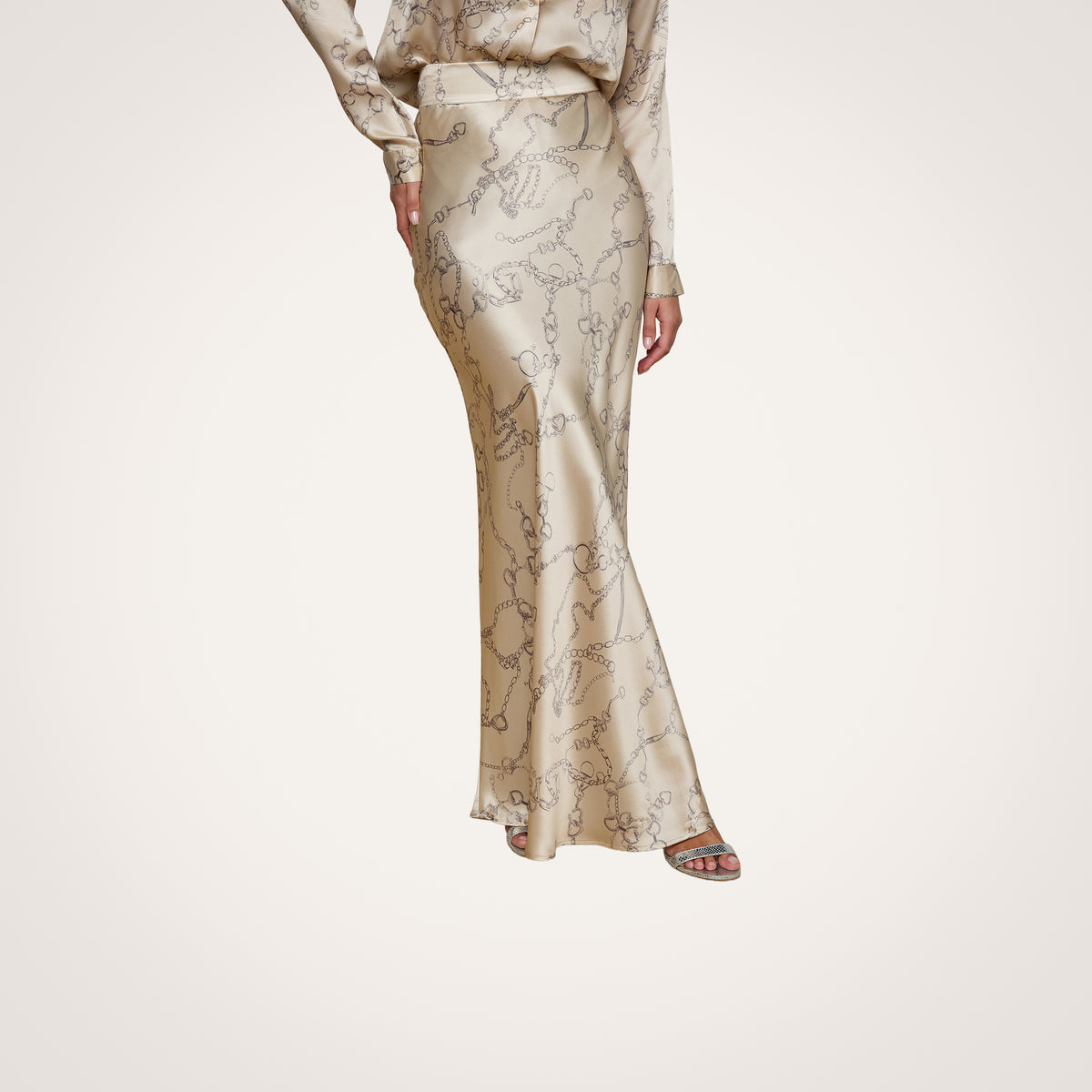 A person wears the Ryker Silk Maxi Skirt in shiny beige with a subtle horsebit chain print, paired with a matching long-sleeve top and open-toe sandals, creating a look of minimalist sophistication on a light wood floor.