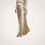 A person embodies minimalist sophistication in the Ryker Silk Maxi Skirt featuring a subtle horsebit chain print, paired with a matching blouse and open-toe sandals, standing on a light wood floor against a plain white wall.