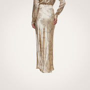 A person wearing the Ryker Silk Maxi Skirt, featuring a long, shiny design with a subtle abstract pattern, stands on a light wood floor facing away from the camera, showcasing minimalist sophistication.