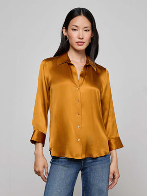 A woman with long dark hair models the L'AGENCE Dani Silk Blouse—a shiny gold, collared top with three-quarter sleeves—paired with blue jeans against a plain light gray background.