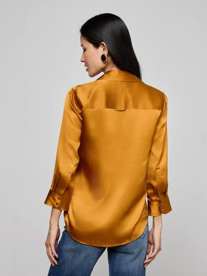 A woman with long dark hair is seen from behind wearing the L'AGENCE Dani Silk Blouse in shiny gold with long sleeves, paired with blue jeans, set against a plain light gray background.
