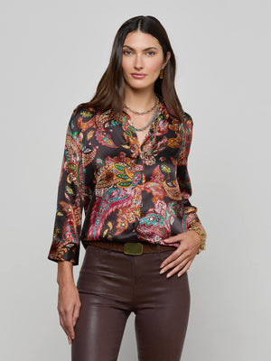 A woman with long brown hair wears the colorful paisley print Dani Silk Blouse by L'AGENCE and dark brown leather pants, accessorized with gold jewelry and a belt, standing against a plain gray background.