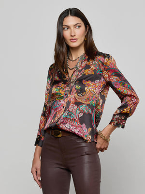 A woman with straight brown hair and gold jewelry stands against a light gray background, wearing the L’AGENCE Dani Silk Blouse with a colorful paisley print and long sleeves, paired with dark brown pants and a neutral expression.