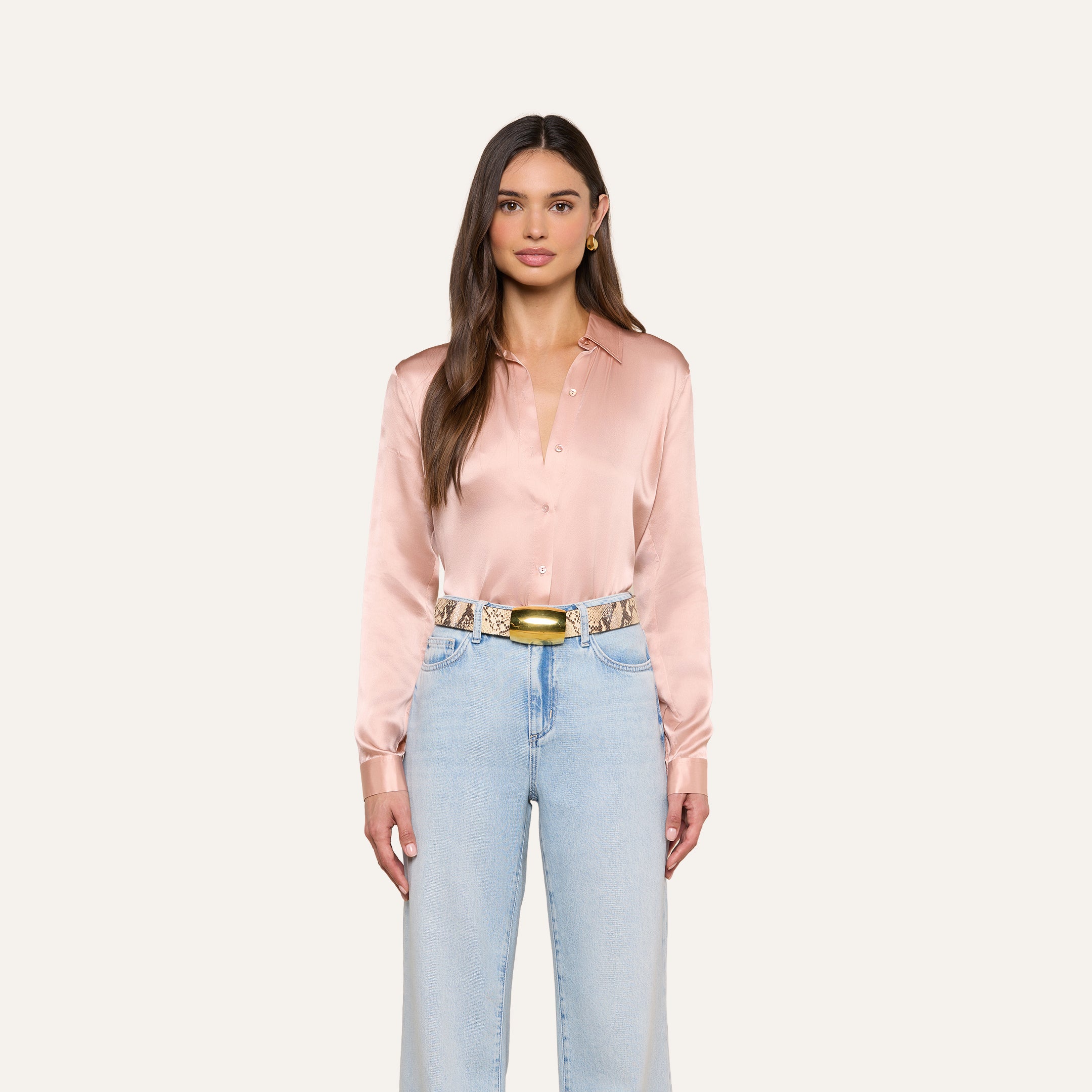 Woman with long brown hair wearing a pink Tyler Silk Blouse by L’AGENCE, light blue high-waisted jeans, a gold belt, and gold hoop earrings, standing against an off-white background.