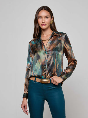 A woman with long brown hair wears the L'AGENCE Tyler Silk Blouse, featuring a colorful feather print, paired with teal pants, a brown belt, gold earrings, and a gold chain necklace against a plain light gray background.