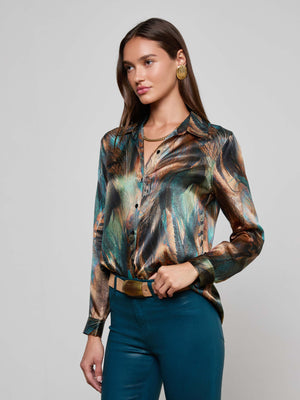 A woman with long brown hair wears the Tyler Silk Blouse by L'AGENCE, featuring a shiny multicolored feather print. She pairs it with teal pants, a gold belt, gold earrings, and a gold necklace, standing against a light gray background.