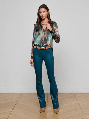 A woman with long brown hair and a neutral expression stands indoors against a white wall, wearing the L'AGENCE Tyler Silk Blouse with feather print in teal, brown, and gold tones, teal fitted pants, tan belt, and tan heels.