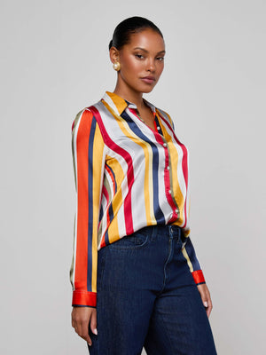 A woman with slicked-back hair and pearl earrings poses against a plain background, wearing the L'AGENCE Tyler Silk Blouse featuring multi-colored stripes in red, yellow, blue, and white, paired with dark blue jeans.