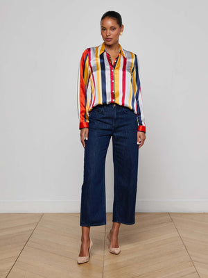 A woman stands indoors against a white wall, wearing the L’AGENCE Tyler Silk Blouse with colorful stripes, paired with dark blue wide-leg jeans and beige heels on a light wood floor.