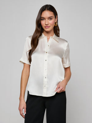A woman with long brown hair smiles softly against a light background, wearing the Ellah Silk Blouse with short sleeves and black pants, her hand casually tucked in her pocket.