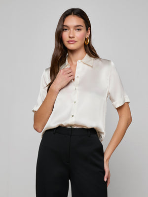 A woman with long brown hair wears the Ellah Silk Blouse, a short-sleeve charmeuse top, tucked into high-waisted black pants against a plain light gray background.