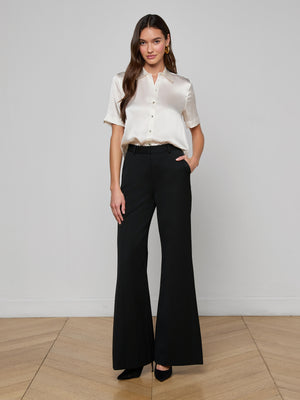 A woman stands against a plain background, smiling slightly, wearing the Ellah Silk Blouse with short sleeves in cream, paired with high-waisted black wide-leg pants and black heels. One hand rests in her pocket.