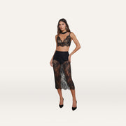 Wearing the Aislin Lace Bralette with adjustable straps and scalloped edges, paired with a matching sheer stretch lace midi skirt and black heels, a woman stands against a plain light background.