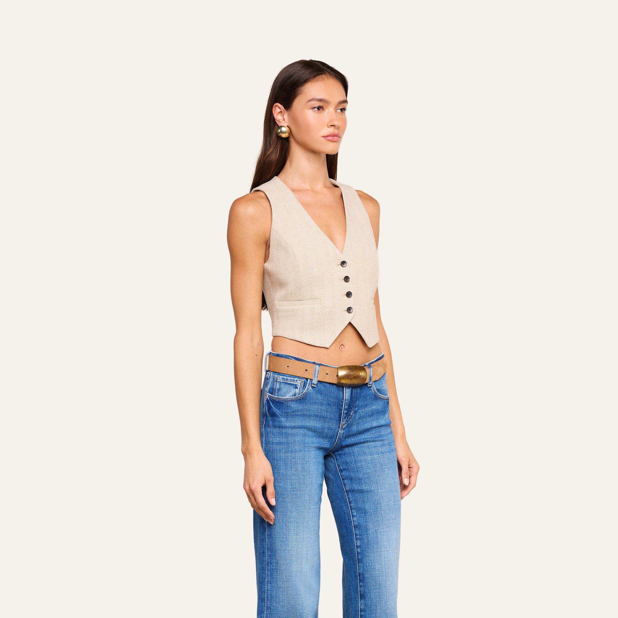 Una donna che indossa il gilet Jianna — un modello senza maniche in tweed beige a spina di pesce con bottoni neri — abbinato a jeans blu con doppia cintura e fibbia dorata, e orecchini dorati, posa su uno sfondo beige a tinta unita, creando un look chic e disinvolto che ricorda la Settimana della Moda di New York.