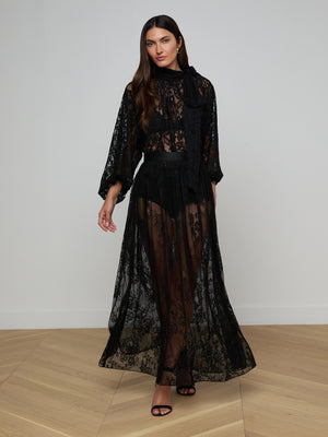 A woman with long brown hair wears the Rhett Oversized Lace Blouse, a sheer black floral lace style with long sleeves, high neck, and bow detail. She stands on a light wood floor against a white wall, wearing black high-heeled sandals.