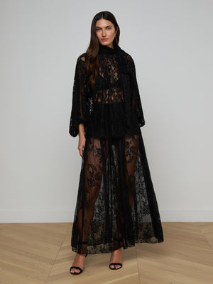 A woman with long, wavy brown hair wears the Rhett Oversized Lace Blouse—sheer black floral lace with wide sleeves—paired with flowing pants and black high-heeled sandals. She stands indoors on light-colored flooring against a plain background.