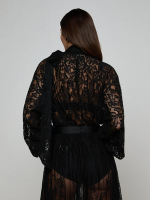 A person with long brown hair faces away, wearing the Rhett Oversized Lace Blouse as a robe with a belt and black undergarments, set against a plain light background.