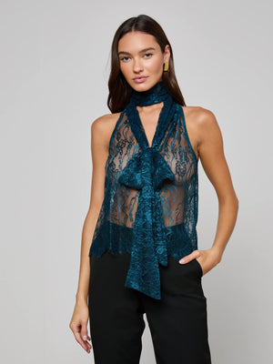 A woman wears the Reece Lace Tie Neck Blouse, a sleeveless teal floral Chantilly lace top with a large bow at the neck, paired with black pants. She stands with one hand in her pocket against a plain light gray background.