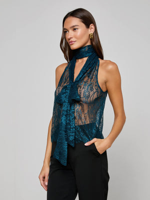 A woman wears the Reece Lace Tie Neck Blouse—sleeveless, sheer teal lace with a matching tie-neck—paired with black trousers. She accessorizes with gold hoop earrings and has long brown hair, one hand casually in her pocket.