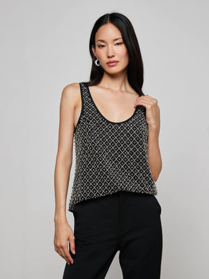 A woman with long dark hair wears the L'AGENCE Ziggy Crystal Tank— a sleeveless black top with a white diamond pattern—paired with black pants. She stands against a light gray background, gazing at the camera neutrally.