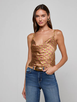 A woman with long brown hair wears the L'AGENCE Calista Silk Camisole in a shiny gold and pink horse bit print, paired with blue jeans and a gold buckle belt, posing with one hand in her pocket against a gray background.