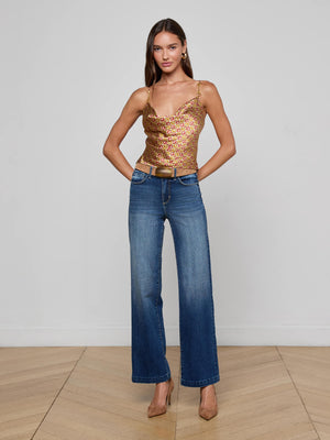 A woman stands on a wooden floor against a white wall, wearing L'AGENCE’s Calista Silk Camisole in a gold and pink horse bit print, blue high-waisted jeans, and tan heels, with her hands in her pockets.