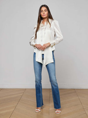 A woman with long brown hair stands indoors on a wooden floor, facing the camera with a neutral expression. She wears the Honey Silk Blouse, blue jeans, and white heeled sandals against a plain backdrop.