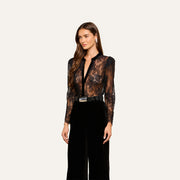 A woman with long brown hair wears the Dorothea Velvet Lace Blouse, paired with black high-waisted pants, a black belt with a silver buckle, and drop earrings, standing against a plain light background.