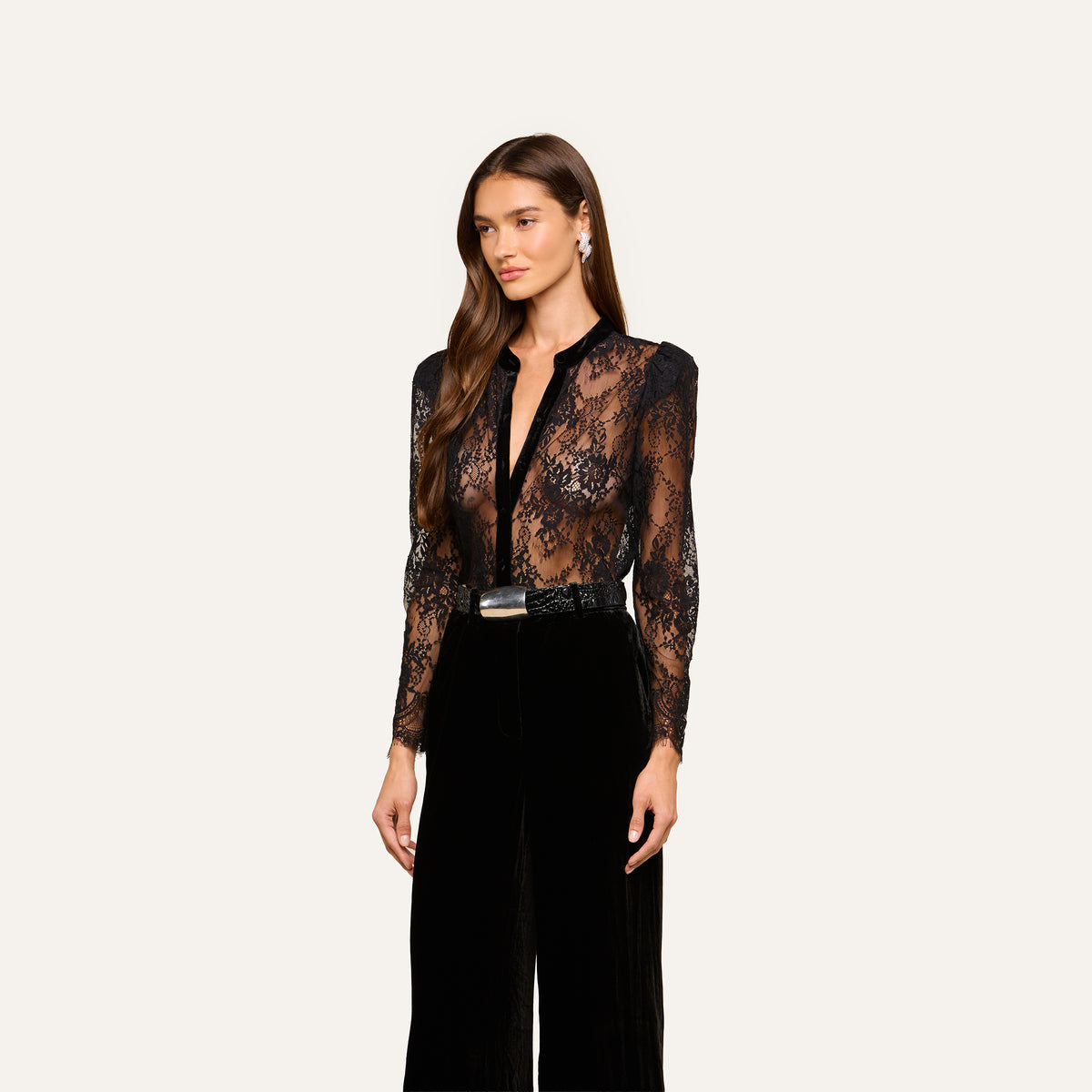 A woman with long brown hair wears the Dorothea Velvet Lace Blouse, paired with black high-waisted pants, a black belt with a silver buckle, and drop earrings, standing against a plain light background.