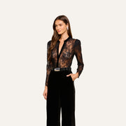 A woman wearing the Dorothea Velvet Lace Blouse—a black long-sleeve top—paired with high-waisted black pants stands against a light background, looking to the side with her hair down.