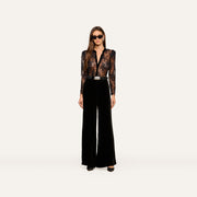 A woman wearing sunglasses, the Dorothea Velvet Lace Blouse—featuring sheer Chantilly lace with velvet seams—and high-waisted black wide-leg pants stands facing forward against a plain white background.