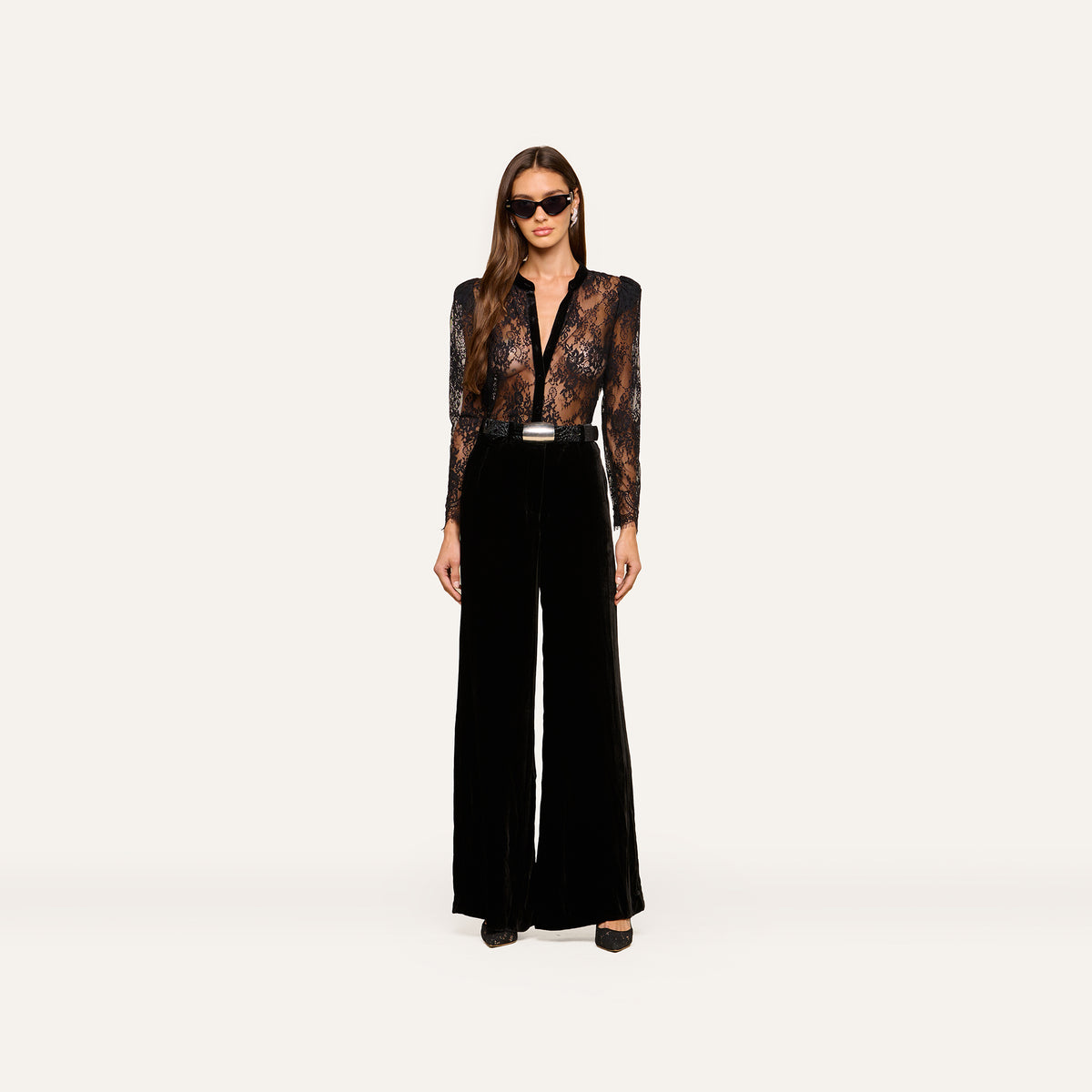 A woman wearing sunglasses, the Dorothea Velvet Lace Blouse—featuring sheer Chantilly lace with velvet seams—and high-waisted black wide-leg pants stands facing forward against a plain white background.