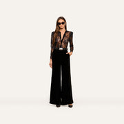 Wearing the Dorothea Velvet Lace Blouse, a woman in sunglasses stands confidently with one hand in her pocket, showcasing its sheer Chantilly lace long sleeves tucked into high-waisted, wide-leg black velvet pants with elegant velvet seams and finished with black shoes.