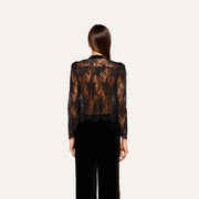 A woman with straight brown hair faces away, wearing the Dorothea Velvet Lace Blouse—crafted from sheer Chantilly lace with long sleeves—and black pants, set against a plain light background.
