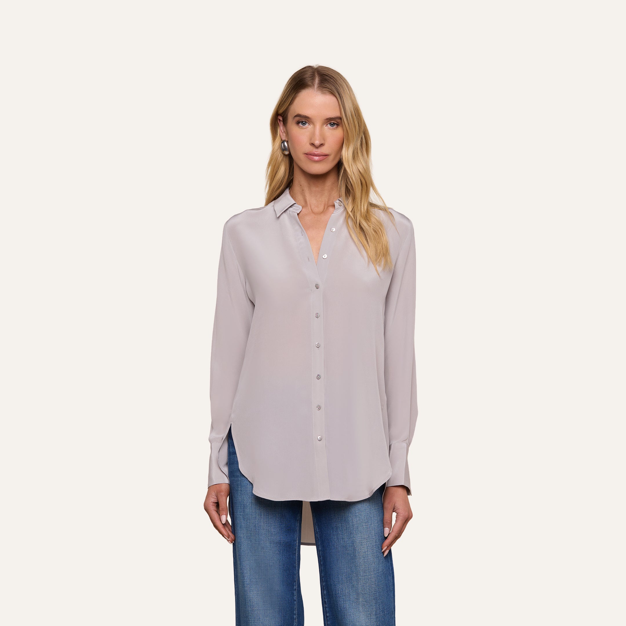 A woman wearing the Babette Silk Blouse, a light gray button-up with long sleeves and a crepe de chine finish, paired with blue jeans, stands facing forward against a plain, light background.