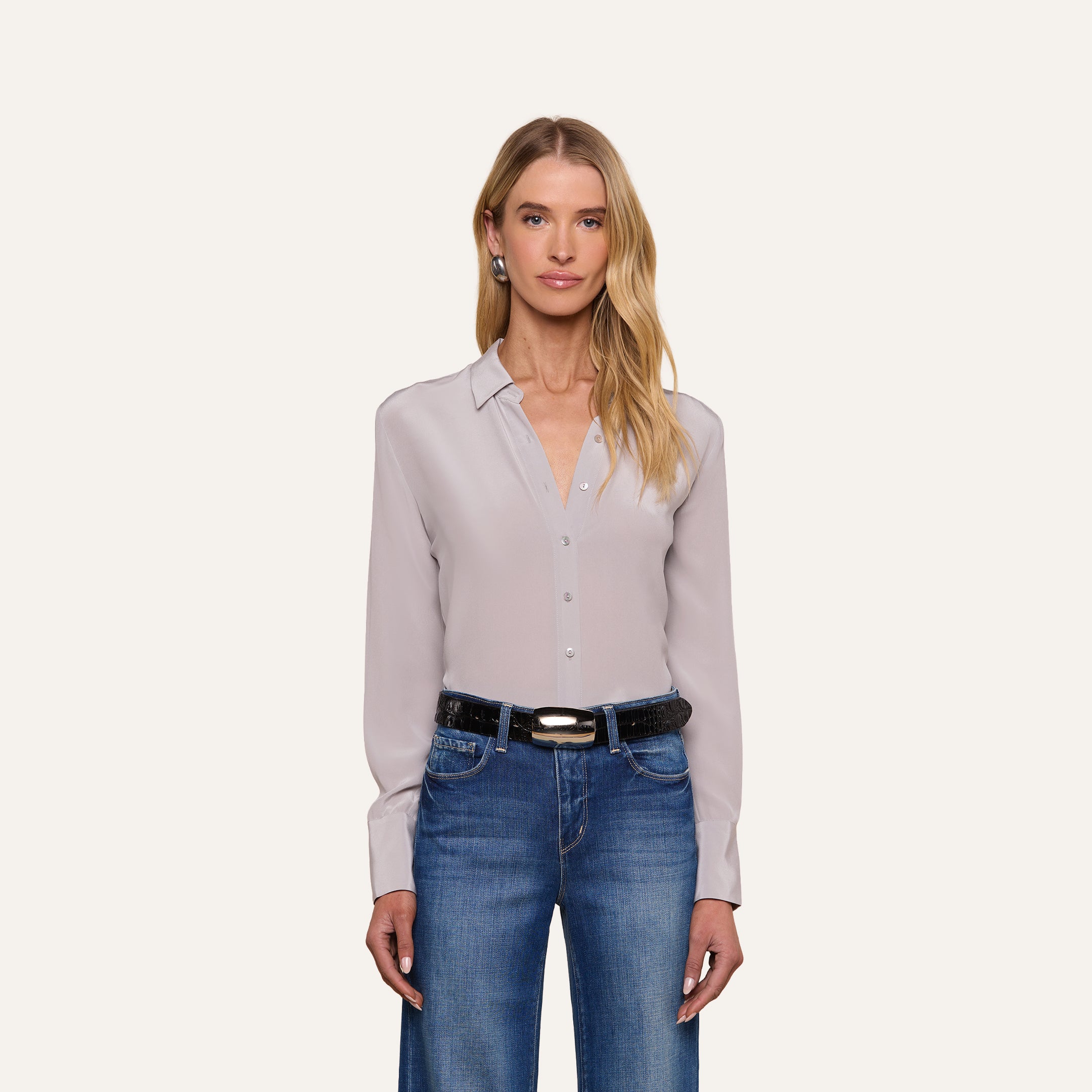 A woman with long blonde hair stands against a plain white background, facing forward with a neutral expression. She is wearing the Babette Silk Blouse in light gray crepe de chine with a relaxed fit, paired with blue jeans.