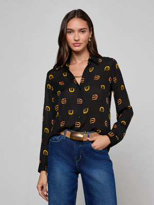 A woman with long brown hair wears the L'AGENCE Nina Silk Blouse—a relaxed-fit black button-down featuring a yellow and orange geometric pattern—tucked into high-waisted blue jeans, posed against a plain light gray background.