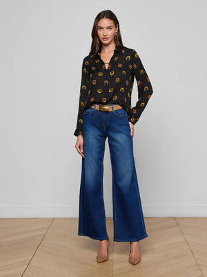 A woman with long brown hair stands indoors against a plain light background wearing the L'AGENCE Nina Silk Blouse, featuring a black buckle print with yellow accents, styled with wide-leg blue jeans, a brown belt, and brown heels.