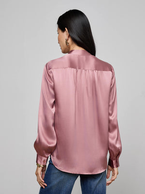 A woman with long dark hair is seen from the back against a plain gray background, wearing L'AGENCE's Bianca Silk Blouse in light pink with long sleeves, paired with blue jeans.