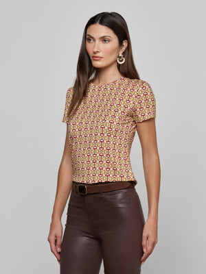 A woman with long brown hair wears the Ressi Fitted Tee in a graphic horse bit print, styled with dark brown pants, a black belt, and large gold hoop earrings, standing against a plain gray background.