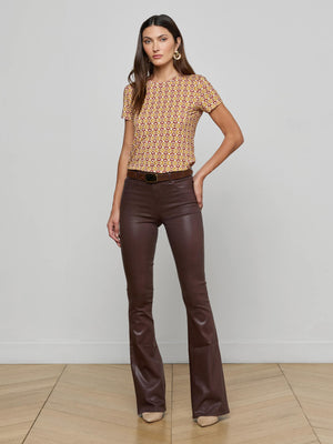 A woman stands indoors on a wooden floor, wearing the Ressi Fitted Tee with a graphic horse bit print, brown flared leather pants with a belt, beige shoes, hoop earrings, and has long hair.