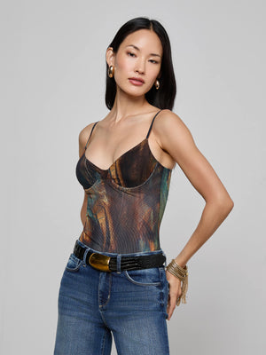 A woman with long dark hair, gold hoop earrings, and gold bracelets wears the L'AGENCE Elowen Mesh Bodysuit with a teal feather print, styled with blue jeans and a black belt, standing against a plain light gray background.
