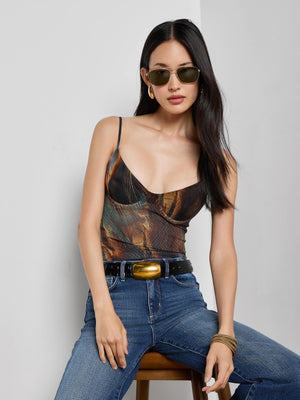 A woman with long dark hair, wearing sunglasses, gold hoop earrings, bracelets, blue jeans, a black belt with a large buckle, and the Elowen Mesh Bodysuit by L'AGENCE sits on a stool against a plain white wall.