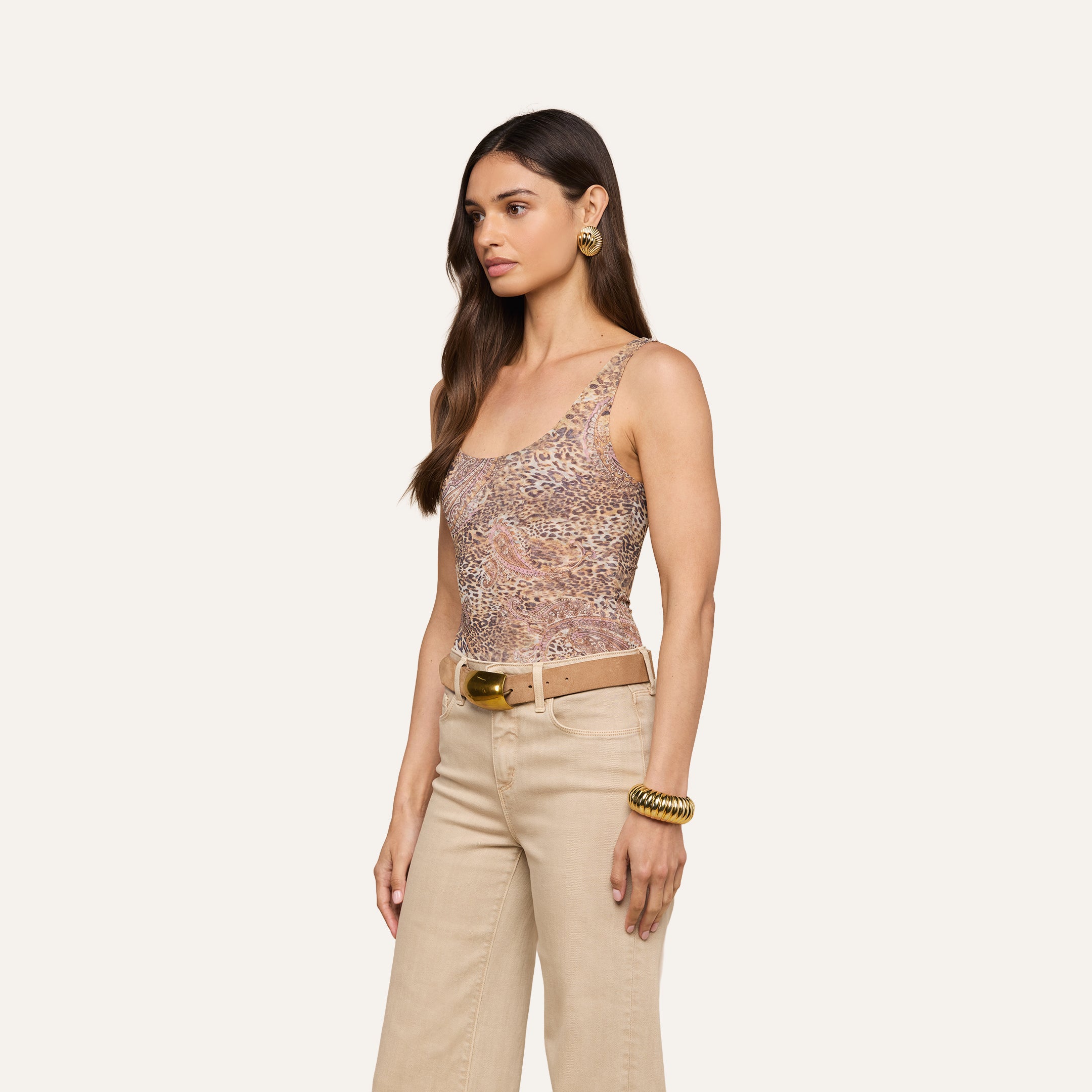 Una donna con lunghi capelli castani indossa il body aderente senza maniche Kalea Mesh, pantaloni a vita alta beige, cintura beige con fibbia dorata, orecchini a cerchio dorati e braccialetti doro su uno sfondo beige.