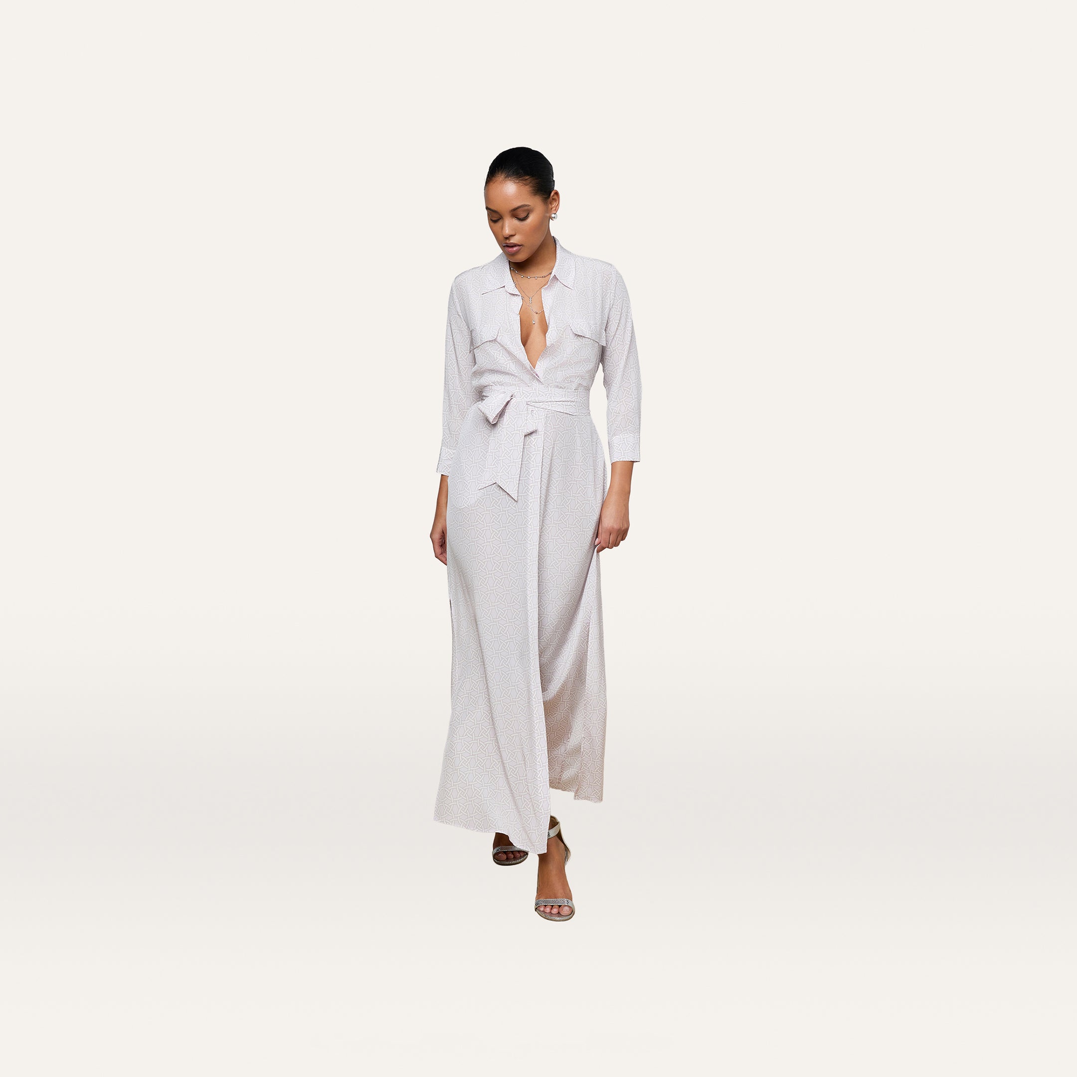 A woman wearing the Cameron Silk Shirt Dress—a long, light-colored crepe de chine wrap dress with a tied waist and three-quarter sleeves—stands against a plain white background, looking down.